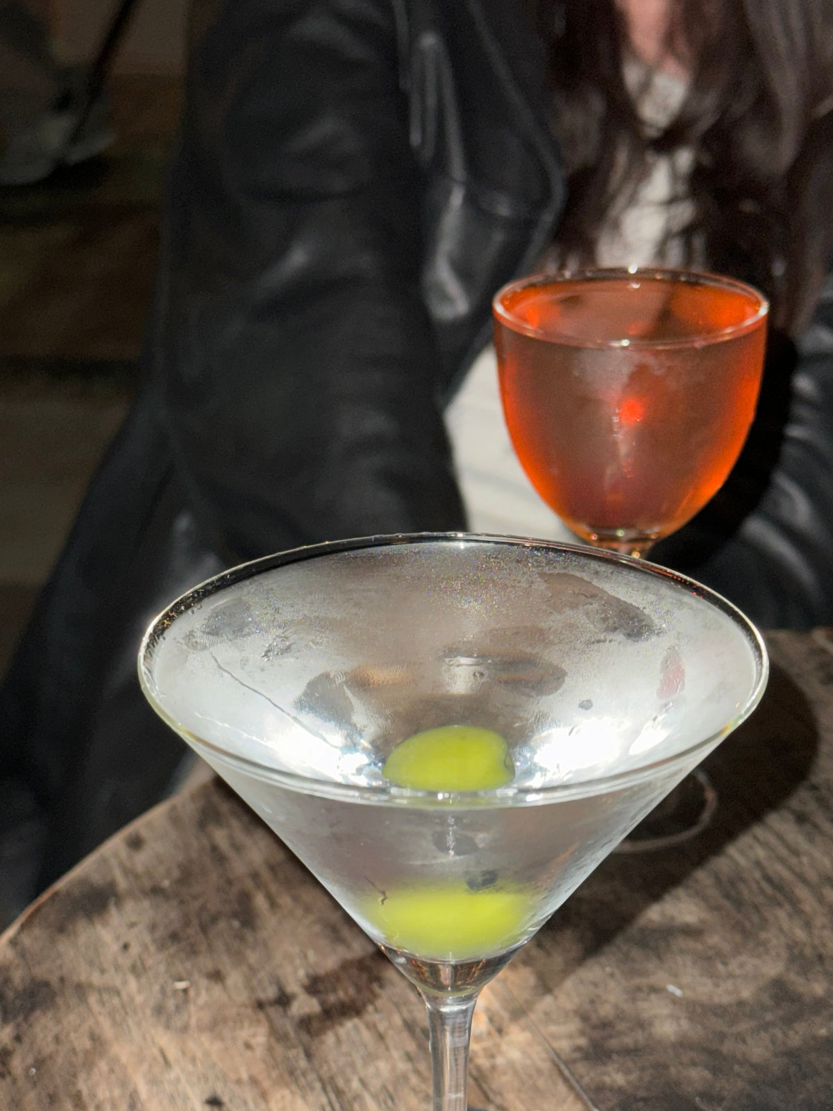Close-up of a cocktail glass with a red liquid and a green olive, blurred background
